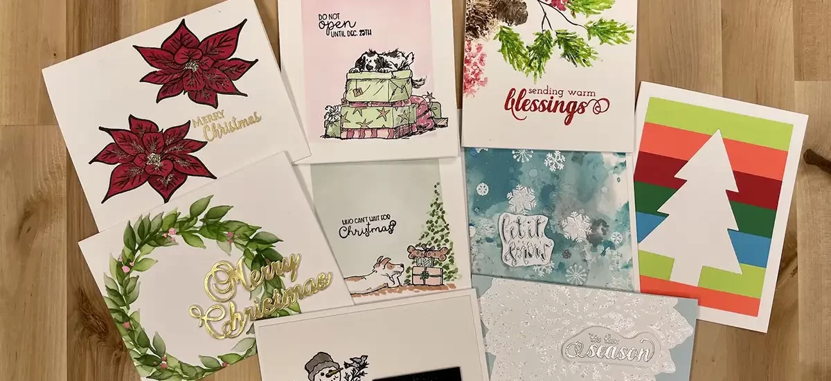 A variety of Christmas greeting cards are displayed on a wooden surface. The designs feature festive elements such as poinsettias, gifts, a snowman, Christmas tree, and seasonal greetings. Learn how to add a personal touch by coloring your favorite Christmas cards.