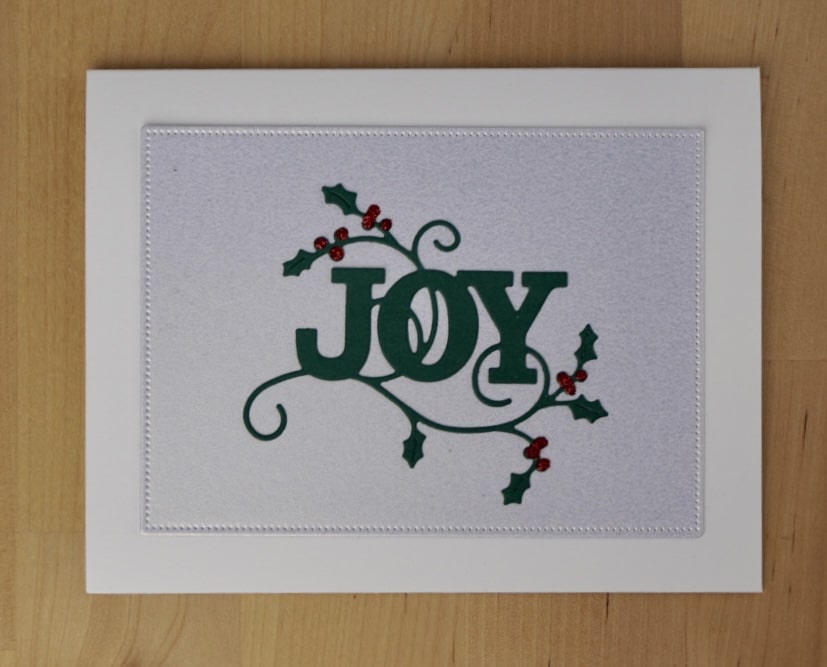 A greeting card with "JOY" in green letters decorated with holly leaves and red berries on a light background. Perfect for those looking to learn how to mass produce cards with festive charm.