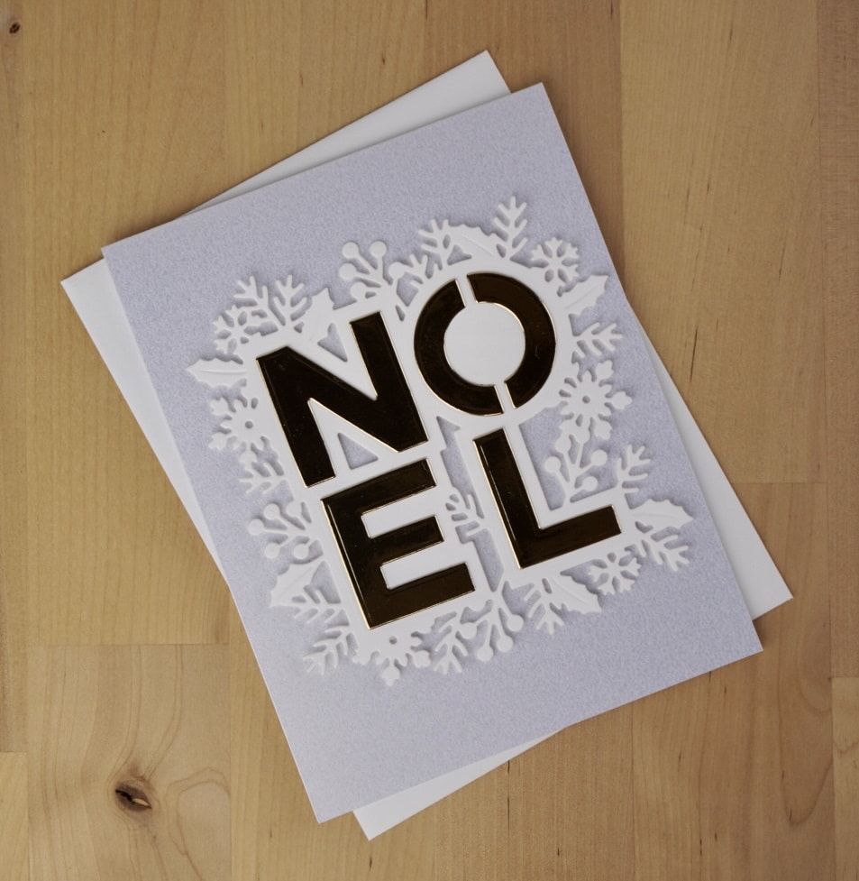 A Christmas card with the word "Noel" in gold letters on the cover, surrounded by white snowflake designs, placed on a light wooden surface. Learn how to mass produce cards like this to share festive cheer with everyone on your list.