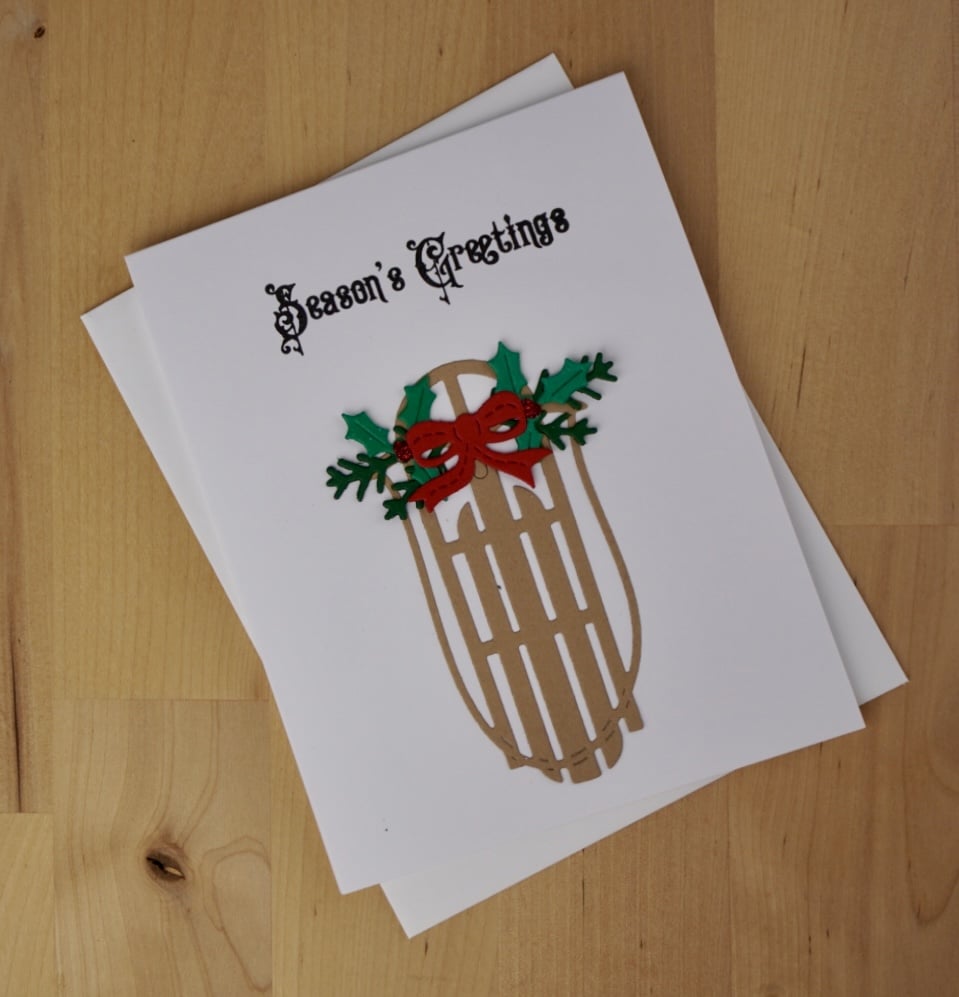 A white holiday card with "Season's Greetings" in black text features an illustration of a wooden sled decorated with holly and a red bow. Perfect for those looking to mass produce cards, this design balances simplicity and festive charm.