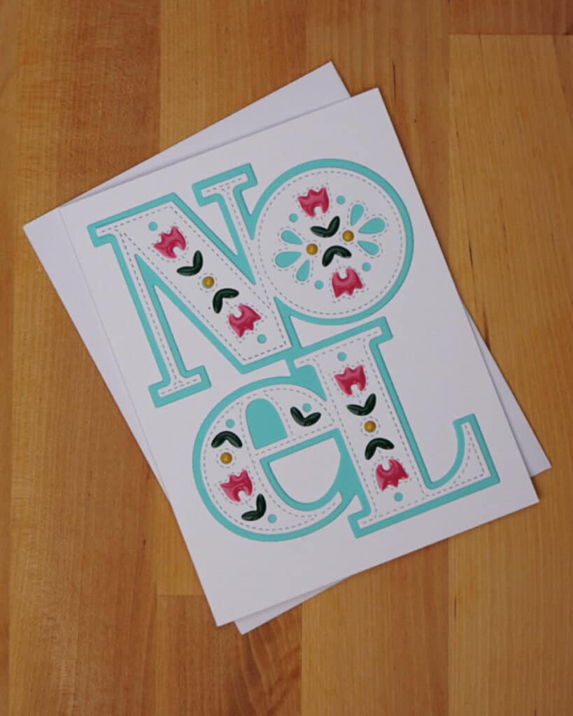 A greeting card on a wooden surface with "NOEL" printed in large, decorative, turquoise letters featuring floral patterns in pink, white, and green. Discover how to make your own Noel Christmas card that catches everyone's eye with its festive charm.