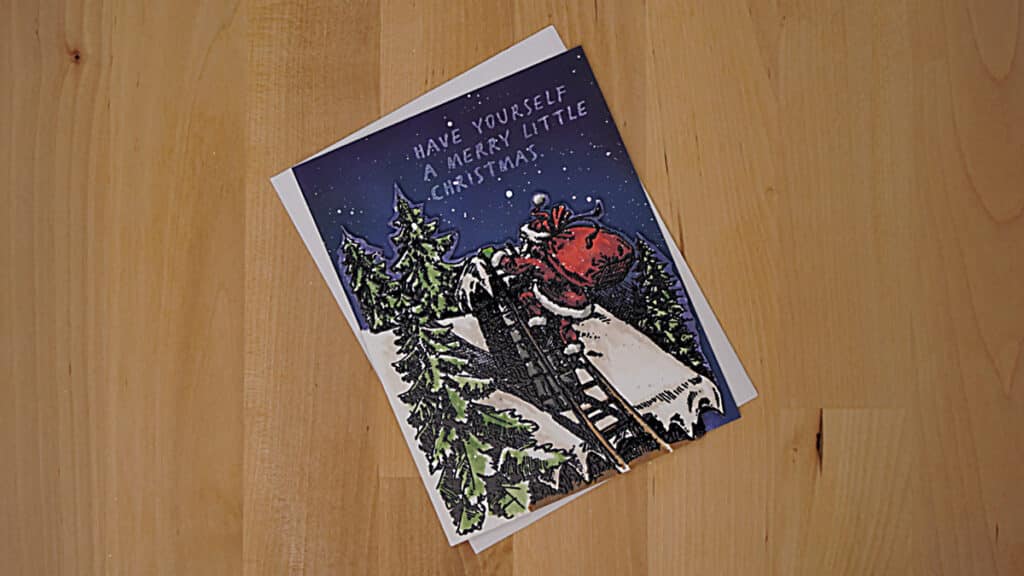 A Christmas card on a wooden surface depicting Santa Claus climbing a ladder onto a snow-covered roof, about to reach the chimney, with the text "Have Yourself A Merry Little Christmas" above.
