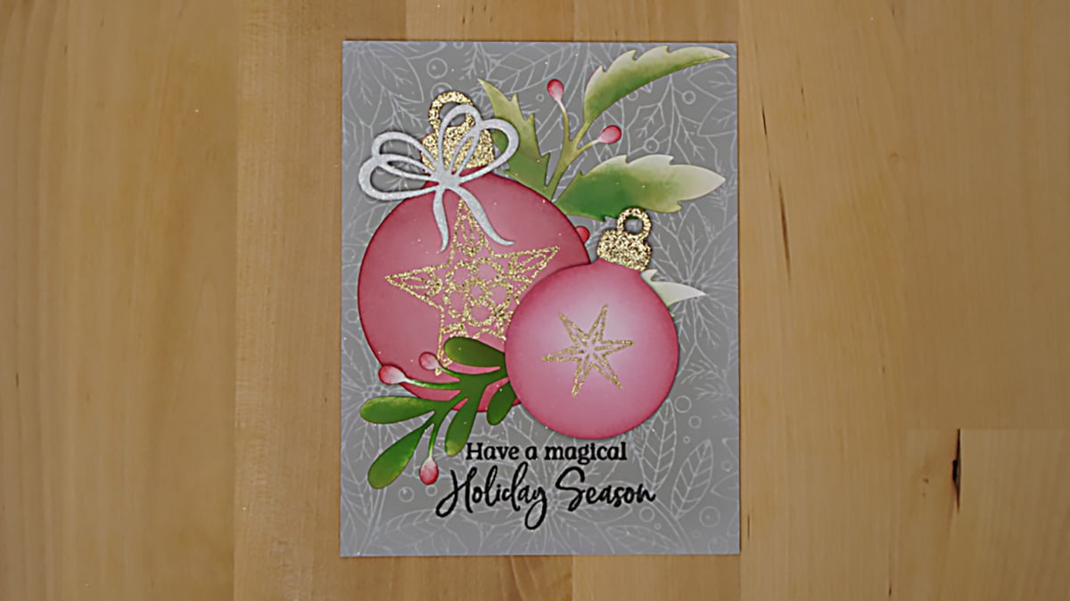 A holiday card featuring pink and gold ornaments, green leaves, berries, and the message "Have a magical Holiday Season" on a gray patterned background. This Blended Ornament Card perfectly captures the festive spirit with its elegant design.