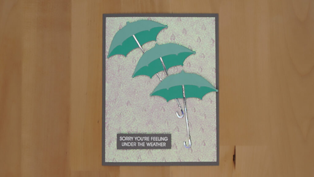 The third of 4 cards made with products from Spellbinders' January holds three teal umbrellas over a glittery raindrop background