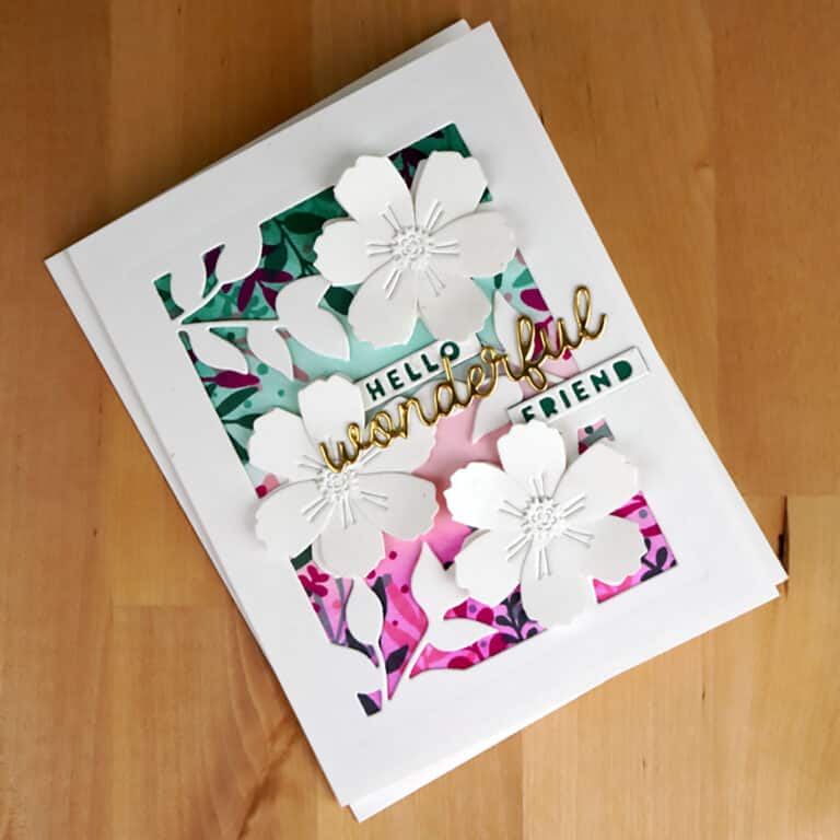 A handmade greeting card featuring white paper flowers, colorful background designs, and the text "Hello wonderful friend" in gold and green lettering. Perfect for sending a touch of joy after a creative day at Cardmaking Summer Camp, the card sits on a wooden surface.