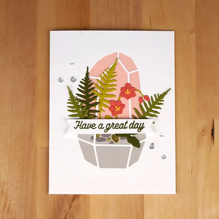 Handcrafted greeting card featuring a geometric planter with green ferns and red flowers, perfect for cardmaking enthusiasts. It has the message "Have a great day" displayed on the front, set against a wooden background.