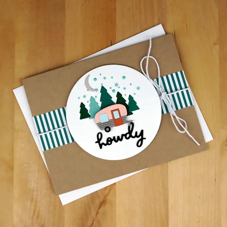 Greeting card on a wooden surface, decorated with a circular design featuring a pink trailer, green trees, a crescent moon, stars, and the word "howdy." A white string is tied around it. This Cardmaking Summer Camp project brings out the fun of summer camp and creativity in cardmaking.