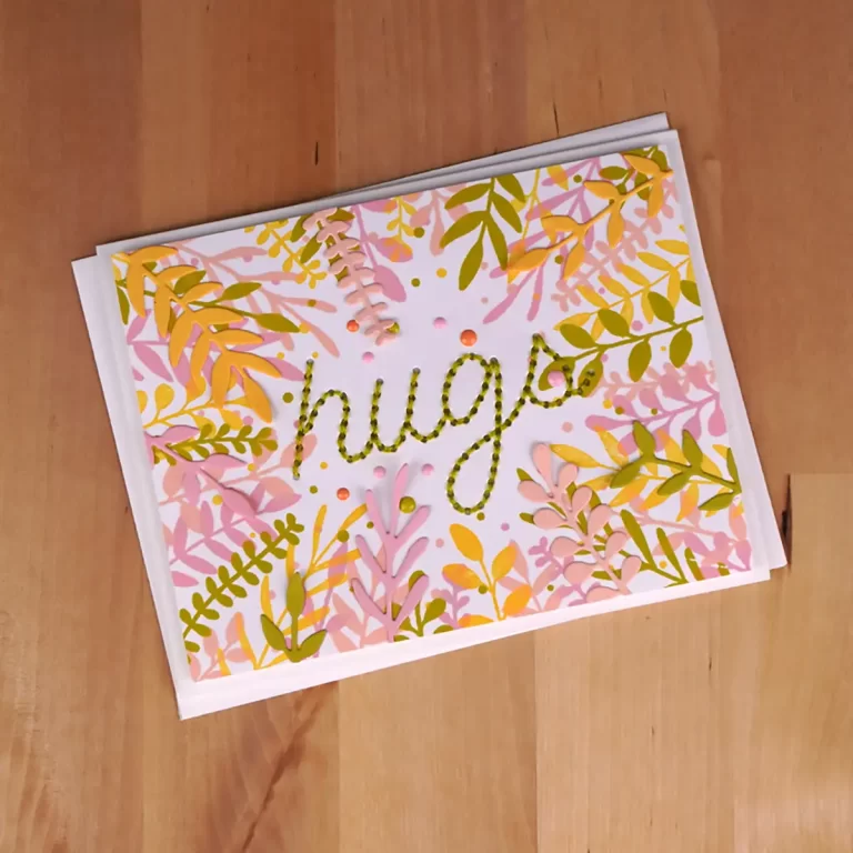 A greeting card with the word "hugs" written in green cursive, surrounded by pink and yellow leaf patterns, lies on a wooden surface, reminiscent of a project from Cardmaking Summer Camp.