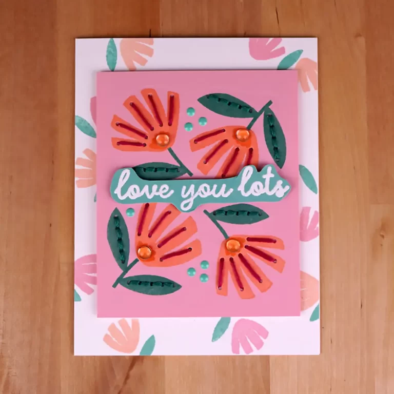 A pink greeting card with layered, floral cutouts and the text "love you lots" in the center, displayed on a floral-patterned envelope on a wooden surface. This charming piece looks like it could have come straight from a summer camp cardmaking session, perfect for sharing heartfelt sentiments.