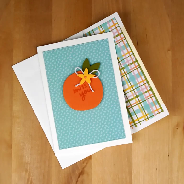 Three greeting cards on a wooden surface. The top card, reminiscent of a vibrant summer camp memory, features a design with an orange fruit and a bow, with the words "miss you" written on it.