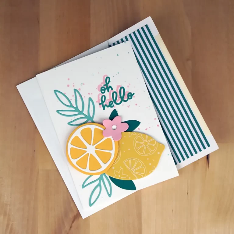 A handmade greeting card with "oh hello" text, featuring a design of a lemon slice, green leaves, and a pink flower, lying on top of an envelope with green stripes—a perfect project for summer camp cardmaking.