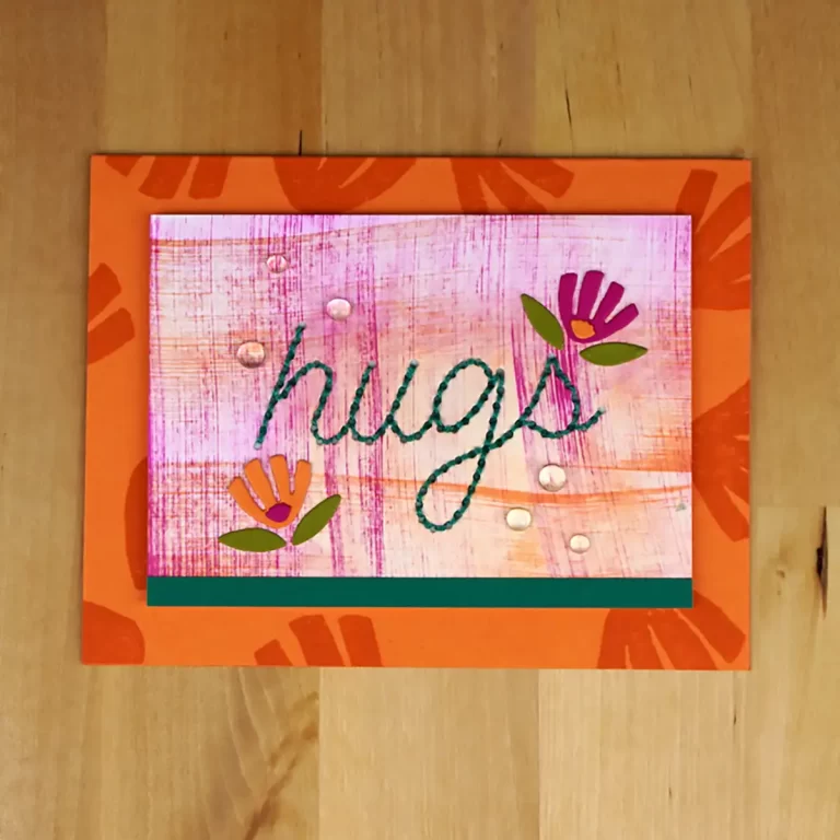 A handmade card on a wooden surface features the word "hugs" stitched in green thread on a pink background with two floral designs and is bordered by an orange frame, showcasing a delightful creation perfect for Cardmaking Summer Camp.