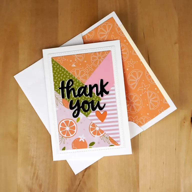 A "thank you" card with a colorful design featuring oranges and patterns, reminiscent of Summer Camp crafts, placed on a wooden surface with a matching envelope nearby.