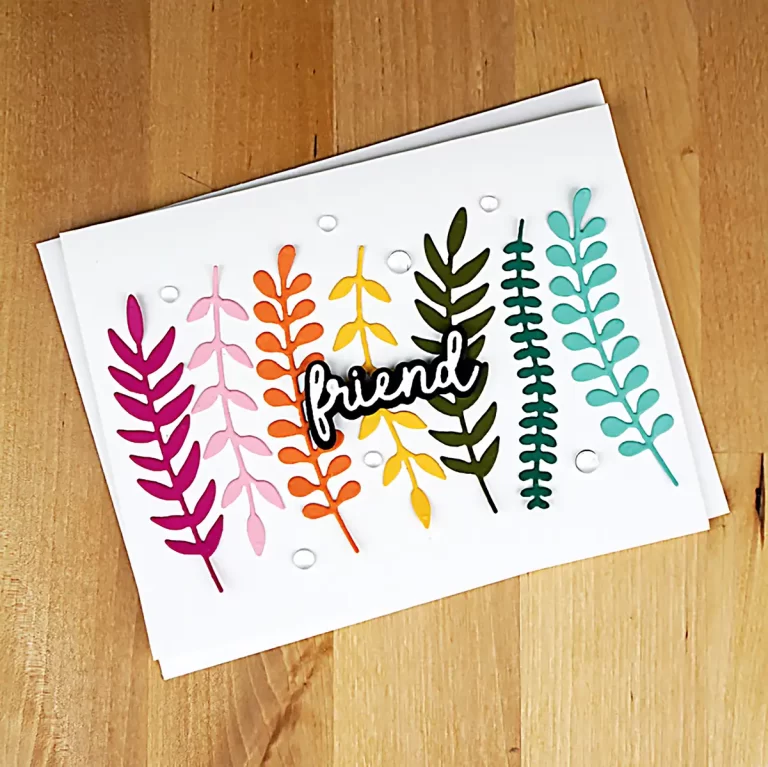 A greeting card with vibrant, paper-cut plant designs and the word "friend" in script at the center. Reminiscent of creations made at a Cardmaking Summer Camp, the card is set on a wooden surface.