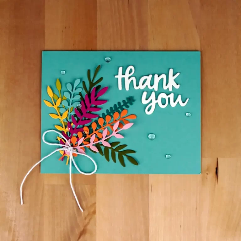 Handmade turquoise thank you card with colorful paper cut-out leaves and white text "thank you," adorned with a white string bow and small clear embellishments, on a wooden surface. Perfect for showcasing your Cardmaking Summer Camp creations.