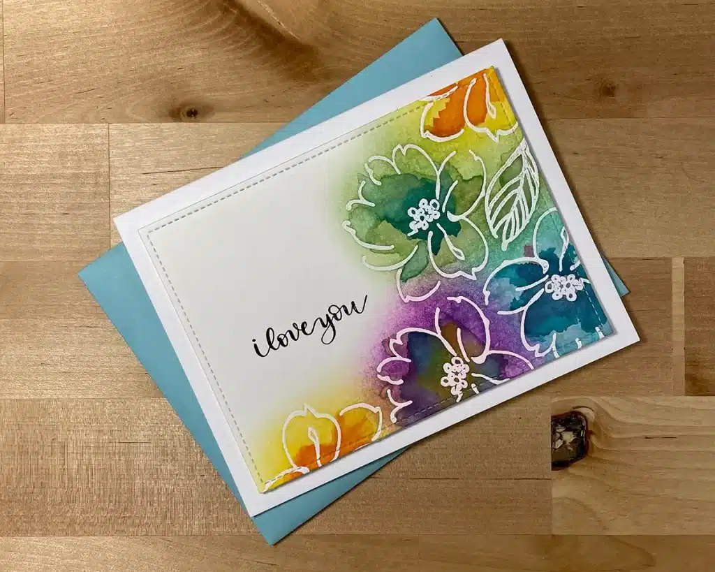 A handmade card with colorful floral watercolor designs, employing a faux watercolor technique, and the handwritten message "I love you" rests on a blue envelope on a wooden surface.
