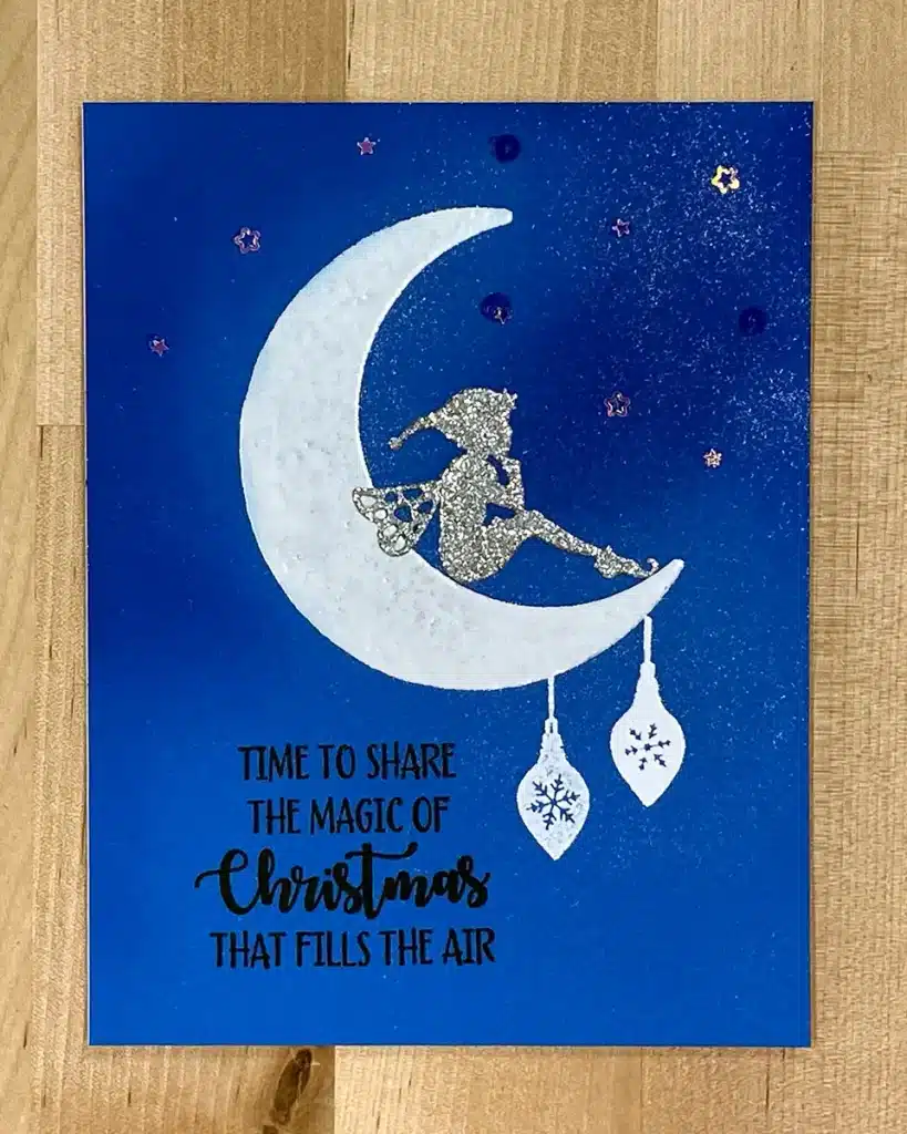 A Christmas card with an illustration of a fairy sitting on a crescent moon, holding gifts. Text reads: "Time to share the magic of Christmas that fills the air." The delicate details are enhanced by heat embossing, making this one of the most enchanting Christmas cards you'll find.