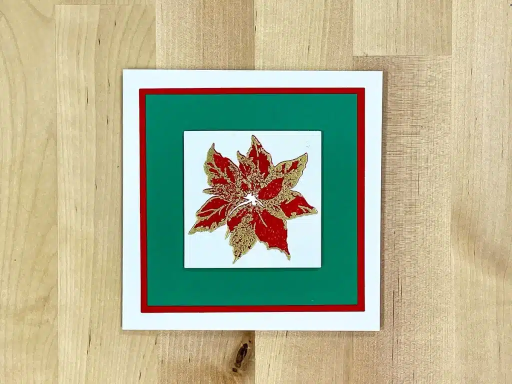 A square, holiday-themed Christmas card with a red and gold poinsettia design in the center, bordered by green and red, placed on a wooden surface. The intricate detailing of the poinsettia is brought to life through exquisite heat embossing.