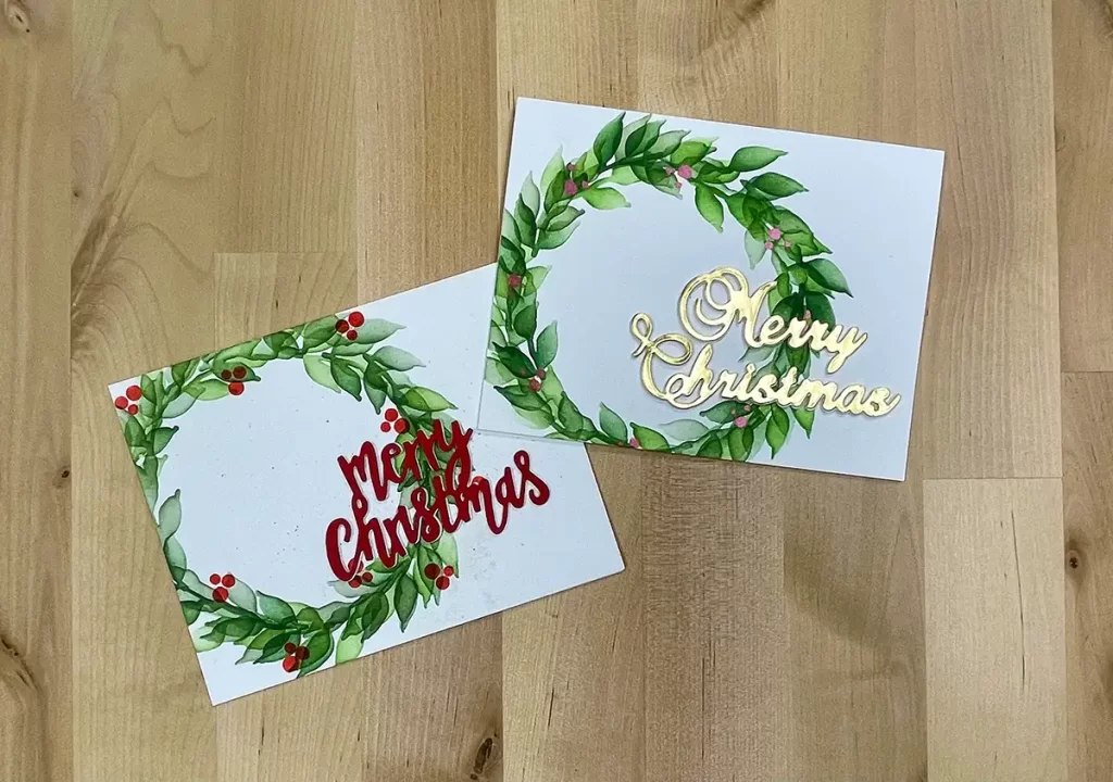 Two color Christmas cards with wreath designs. One features "Merry Christmas" in gold, while the other showcases "Merry Christmas" in red. Both cards are laid on a wooden surface.