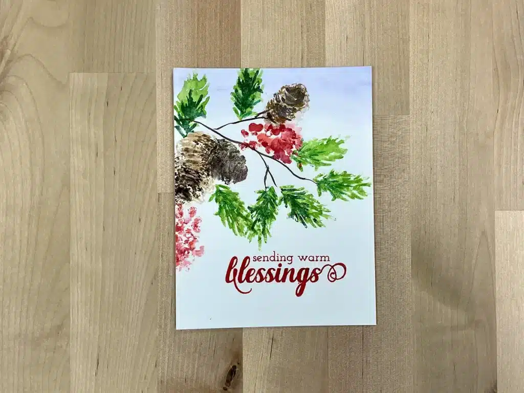 A greeting card featuring a watercolor illustration of pine cones, green leaves, and red berries. The red text on the card reads "sending warm blessings." Perfect for those who love Christmas crafts, this charming Christmas card rests elegantly on a wooden surface.