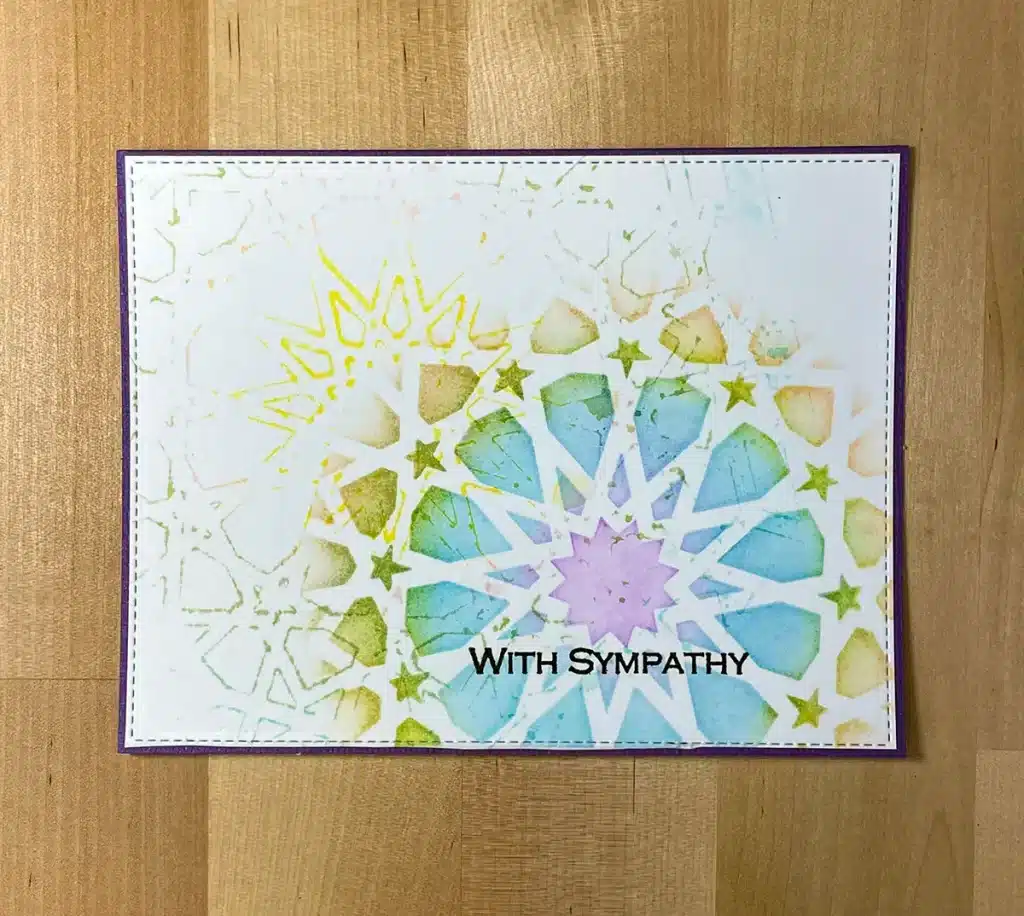 A sympathy card featuring a colorful, geometric pattern with a starburst design on a wooden background. The text "With Sympathy" is printed on the card. Enhance your creations with stencils to easily replicate this beautiful design or explore making 5 cards in various styles.