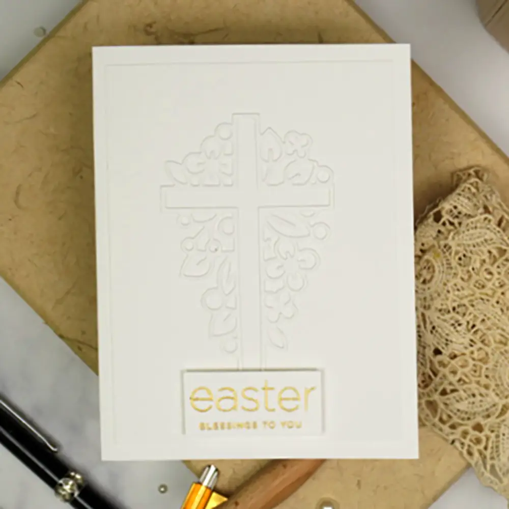 White Easter greeting card with an embossed cross and floral design, featuring the text "Easter Blessings to You" in gold. The card lies on a clipboard next to a fountain pen and a piece of lace ribbon.