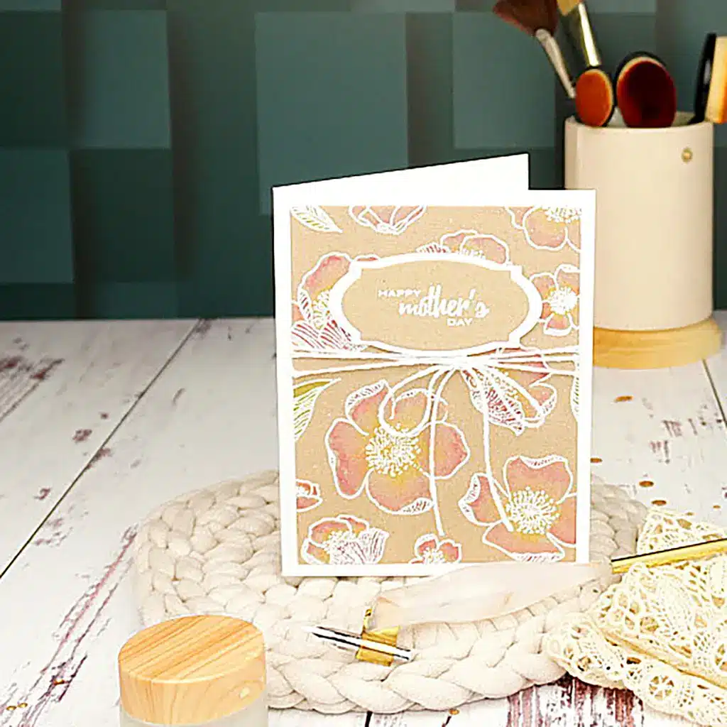A Mother's Day card with a floral design is placed on a crocheted mat, surrounded by art supplies including paintbrushes in a container and small wooden items, as part of the Altenew Celebrating Parents Card Drive on a wooden table.