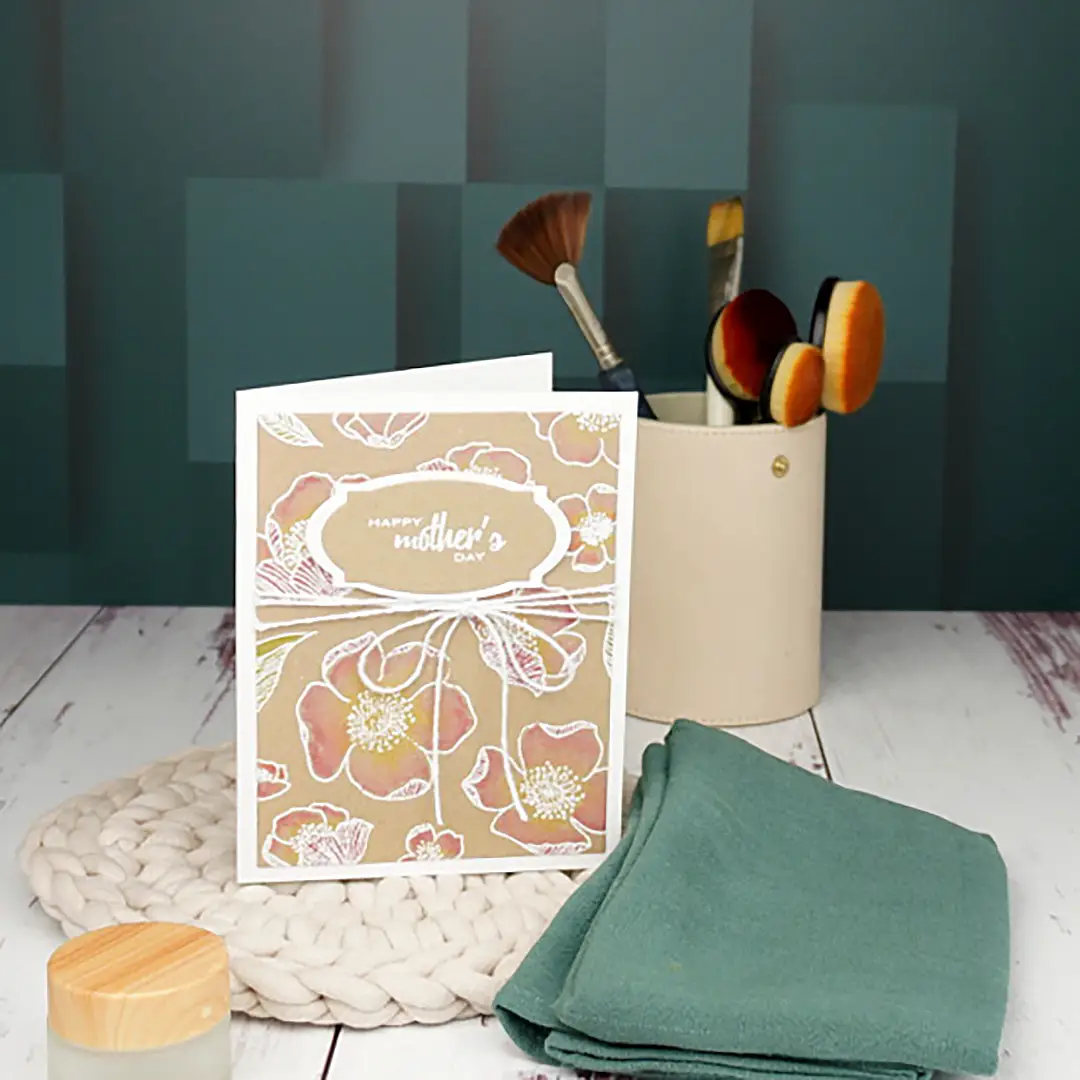 A Mother's Day card with a floral design, featuring Altenew, stands on a white wooden surface beside make-up brushes in a beige holder, a folded green cloth, and a woven mat.