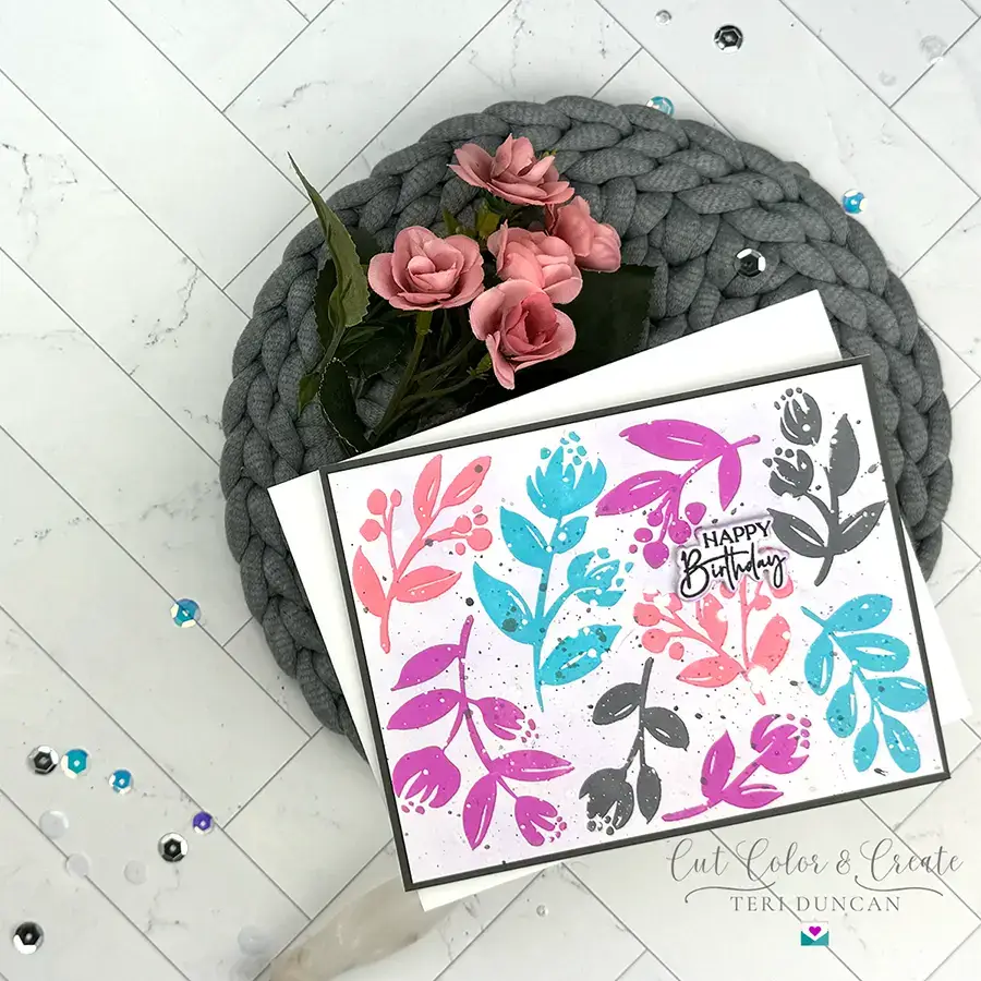A colorful birthday card adorned with sprigs and floral patterns in pink, blue, and gray lies on a gray woven placemat, accompanied by a bouquet of pink roses on a white tiled surface. Confetti is scattered around.