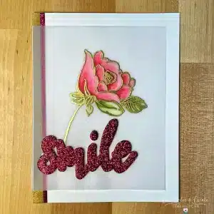 A handmade greeting card featuring a watercolor pink rose and the word "Rosey Smile" written in glittery pink letters on a transparent overlay. The card has gold and pink glitter borders on the left edge.