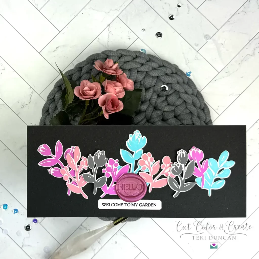 A handmade card with colorful floral designs and a wax seal saying "HELLO". Below, a label reads "WELCOME TO MY GARDEN." The card is beautifully set on a grey textured mat adorned with pink roses.