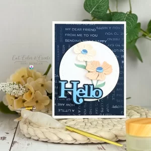 A handmade greeting card with a blue "Hello" in the center, floral decorations, and a background featuring repeated text. The Hello Card is displayed on a woven mat with decorative items nearby.