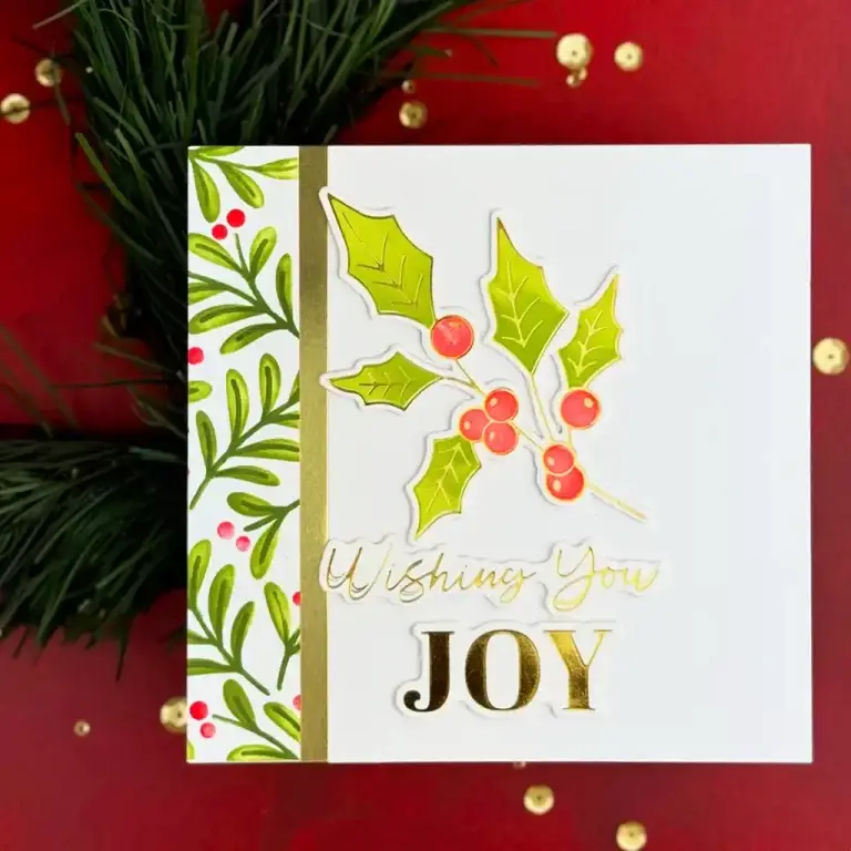 A holiday greeting card designed with Spellbinders July 24 Monthly kit with holly and berries on the front and the phrase "Wishing You Joy" in gold letters. The card is set against a red background with greenery and golden beads, perfect for sending cheer in July 2024 or gifting through Monthly Clubs.