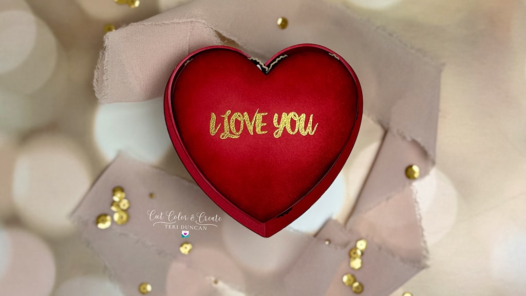 Heart-shaped box of chocolates adorned with "I LOVE YOU" in gold text, rests atop a torn piece of paper, surrounded by scattered gold sequins.