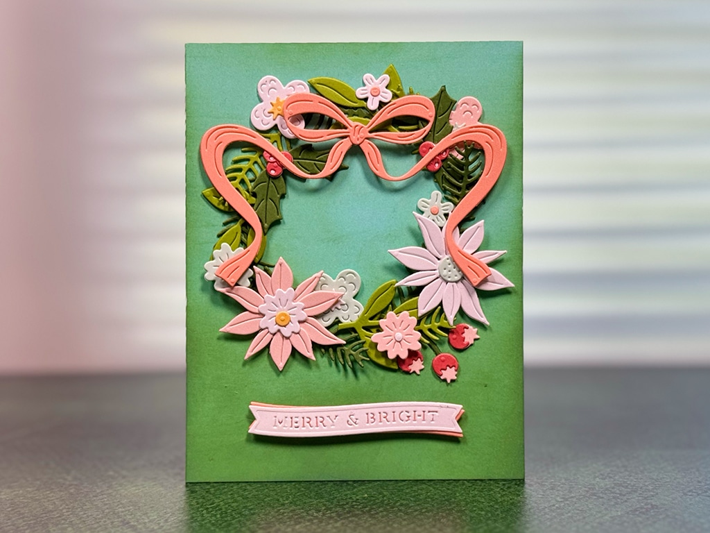 A green greeting card featuring a decorative floral wreath with pink flowers and a ribbon, and a "Merry & Bright" label at the bottom. Created with the Gifting Bow Set from Spellbinders Merry Moments Collection.