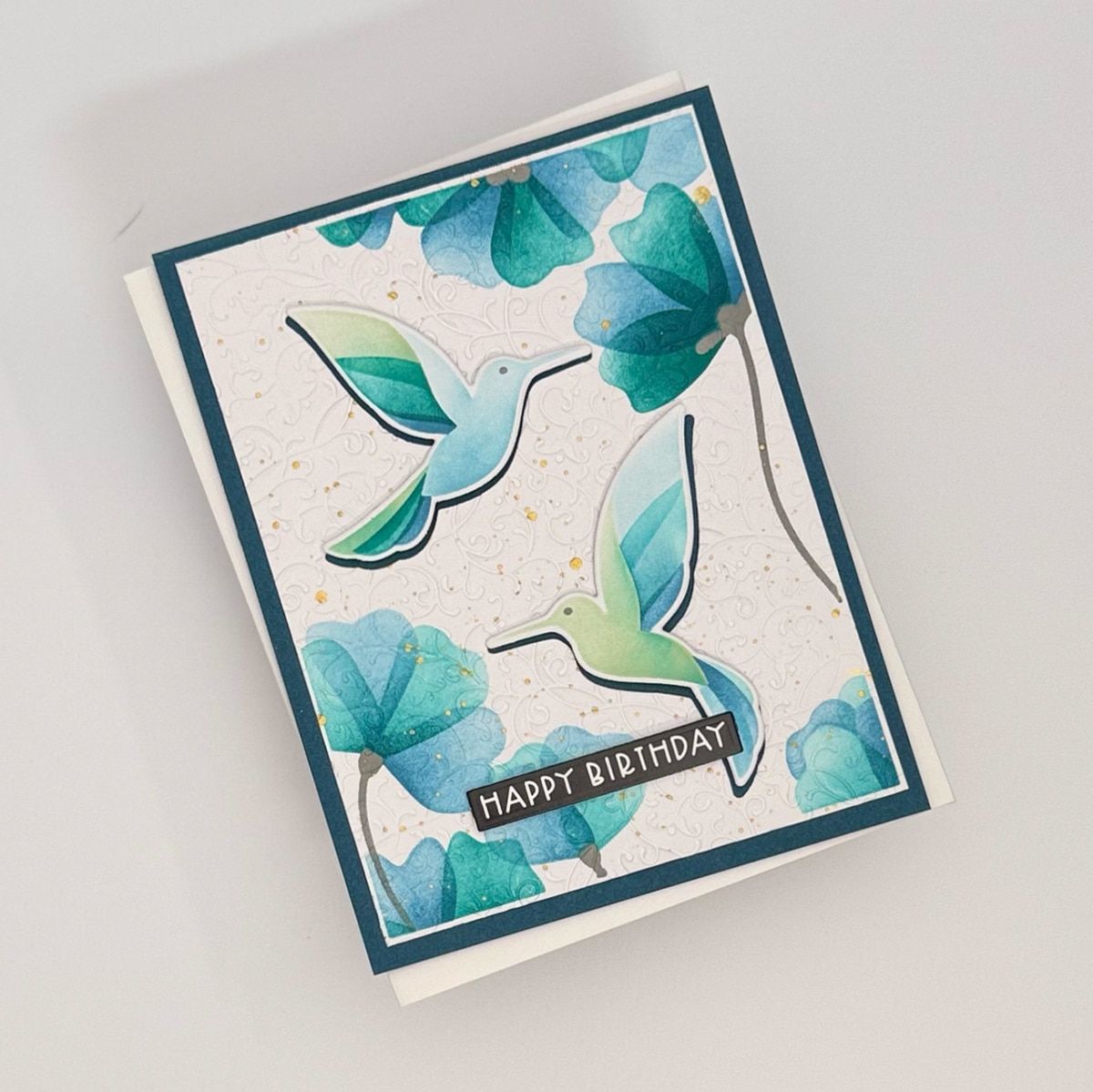 A set of 4 handmade cards featuring two illustrated hummingbirds and blue-green flowers, with "Happy Birthday" written on a black and white label. Part of the charming Flit & Flitter collection.