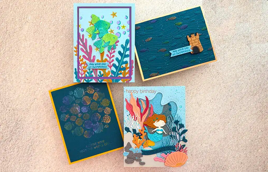 Four handmade Fish Cards with ocean-themed designs—including fish, a castle, sea plants, and a mermaid—from the August 2025 Kits collection are displayed on a beige surface.