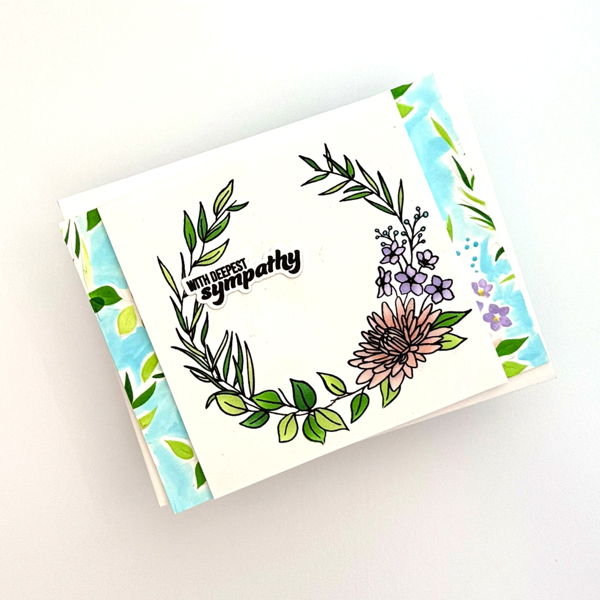 The Floral Deepest Sympathy card featuring a hand-drawn floral wreath and "With deepest sympathy" text, adorned with delicate flowers and leaves on a white background with blue and green borders.