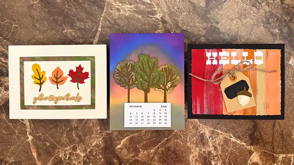 Three autumn-themed handmade cards are displayed on a brown marble surface, featuring leaves, a calendar, and a "HELLO" greeting. Cards created using Spellbinders Autumn Archives Collection.