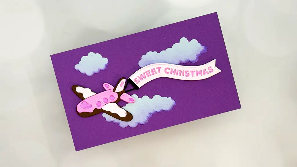 A handmade card from the Candy Christmas Collection features a pink airplane pulling a "Sweet Christmas" banner, flying among clouds on a purple background—perfect for unique Christmas card ideas.