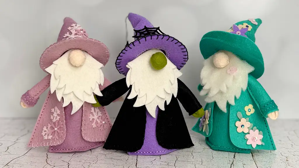 Three felt gnome figurines dressed as wizards in pink, purple, and teal outfits, each with a long white beard and decorated hats, stand on a light textured surface. Created using Spellbinders 3D Felt Collection