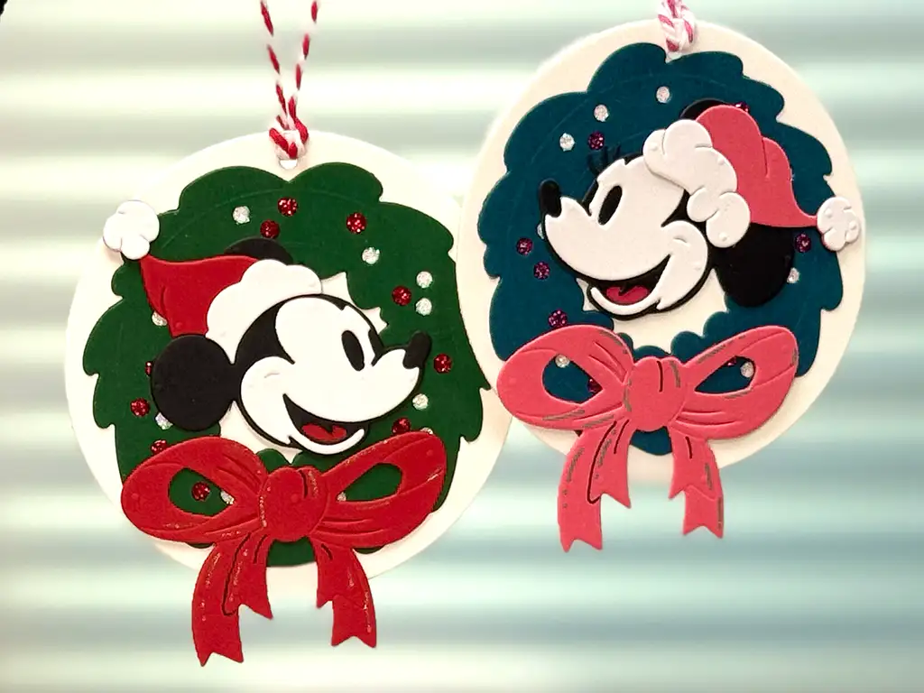 Two round Christmas ornaments featuring cartoon mouse faces, each wearing a Santa hat, with wreath designs and red bows, hanging on striped string.