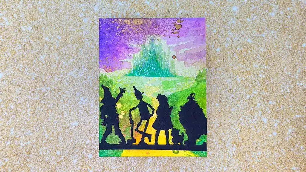 A colorful illustration shows four silhouetted figures and a dog walking toward a green city on the horizon under a purple and yellow sky. This card was created using products from Spellbinders Wonder of Oz Collection.