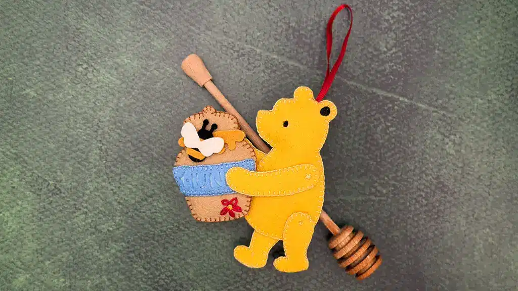 A felt ornament of a yellow bear holding a blue honey pot with bees, attached to a wooden honey dipper with a red hanging loop, on a textured green background.