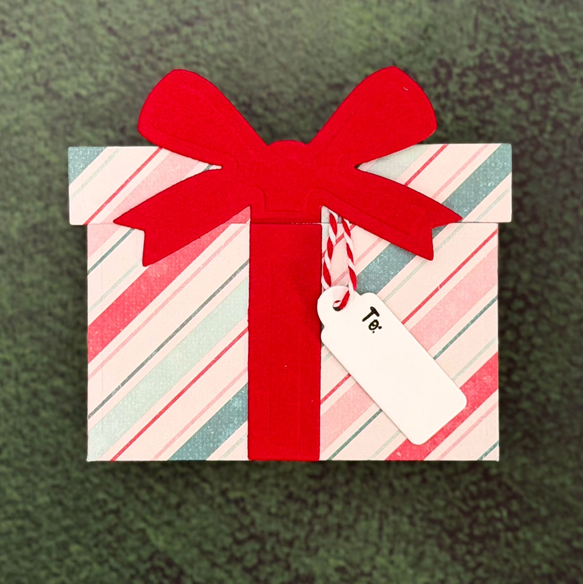 A rectangular Christmas gift box with red and pastel diagonal stripes, topped with a large red bow and a white tag labeled “To:”, sits on a green textured surface. Perfect as a festive Christmas package or gift card holder.