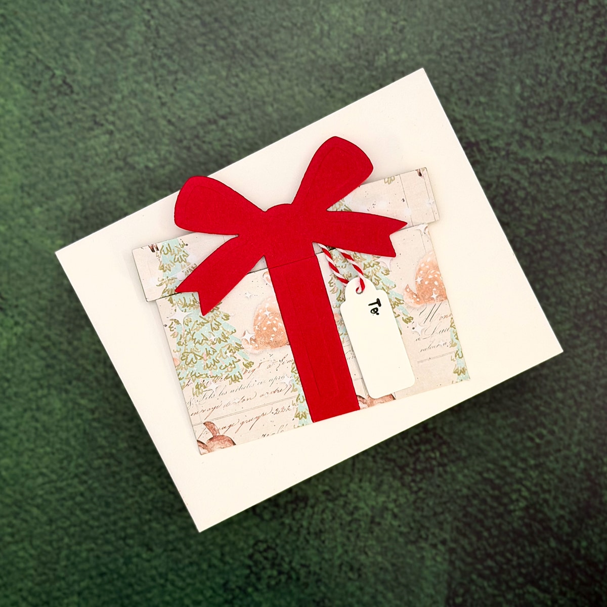 A white greeting card featuring a cut-out present design with a red bow and small gift tag, perfect as a Christmas package or festive gift card holder, placed on a textured green surface.