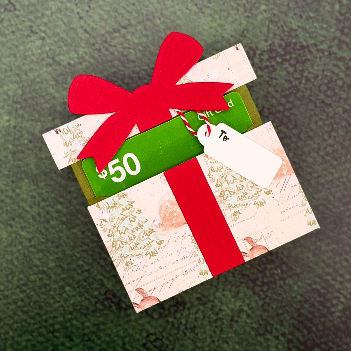 A $50 gift card in a festive envelope, decorated with a red bow and gift tag, makes the perfect Christmas Gift as it rests on a textured green surface.