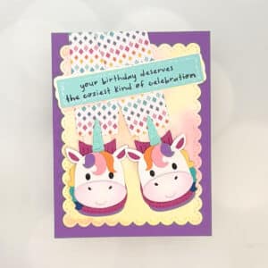 A Unicorn Birthday Card featuring two cartoon unicorn slippers and a sign that reads, "your birthday deserves the coziest kind of celebration," set against a playful purple and yellow background.
