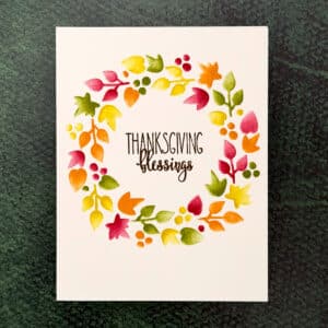 A Thanksgiving Card featuring "THANKSGIVING blessings" at the center, encircled by a colorful Blessings Wreath of painted autumn leaves and berries on a green background.