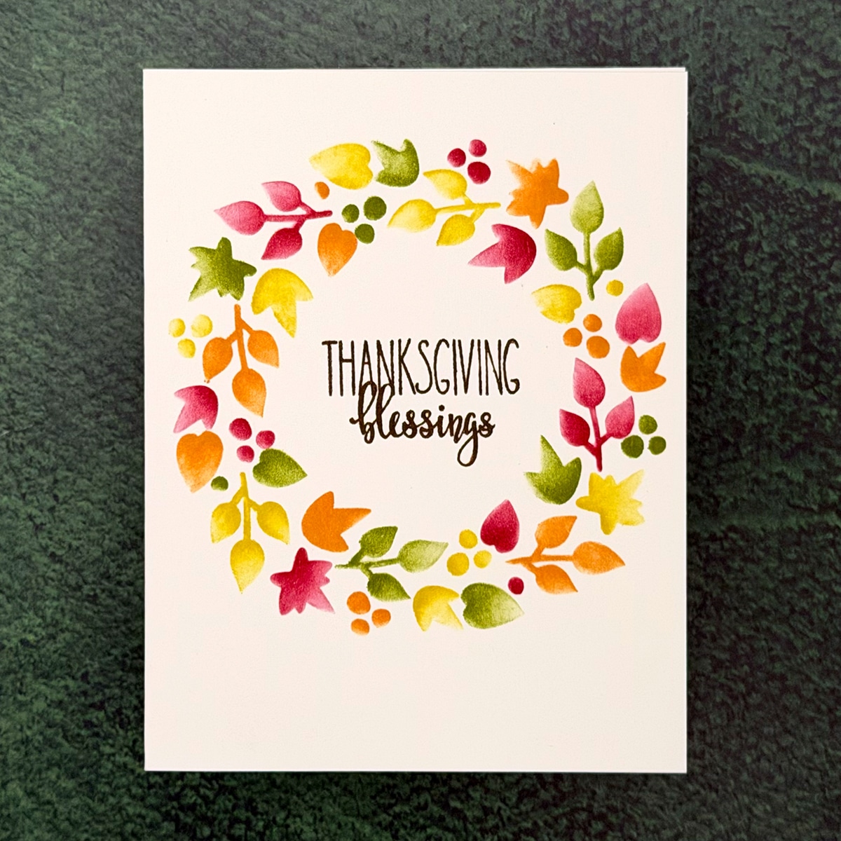 A Thanksgiving Card featuring "THANKSGIVING blessings" at the center, encircled by a colorful Blessings Wreath of painted autumn leaves and berries on a green background.